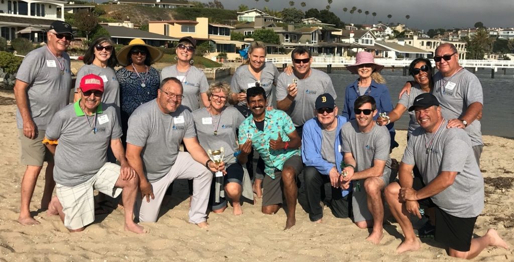 Twenty adults of different backgrounds, genders, and ages pose together on a beach.